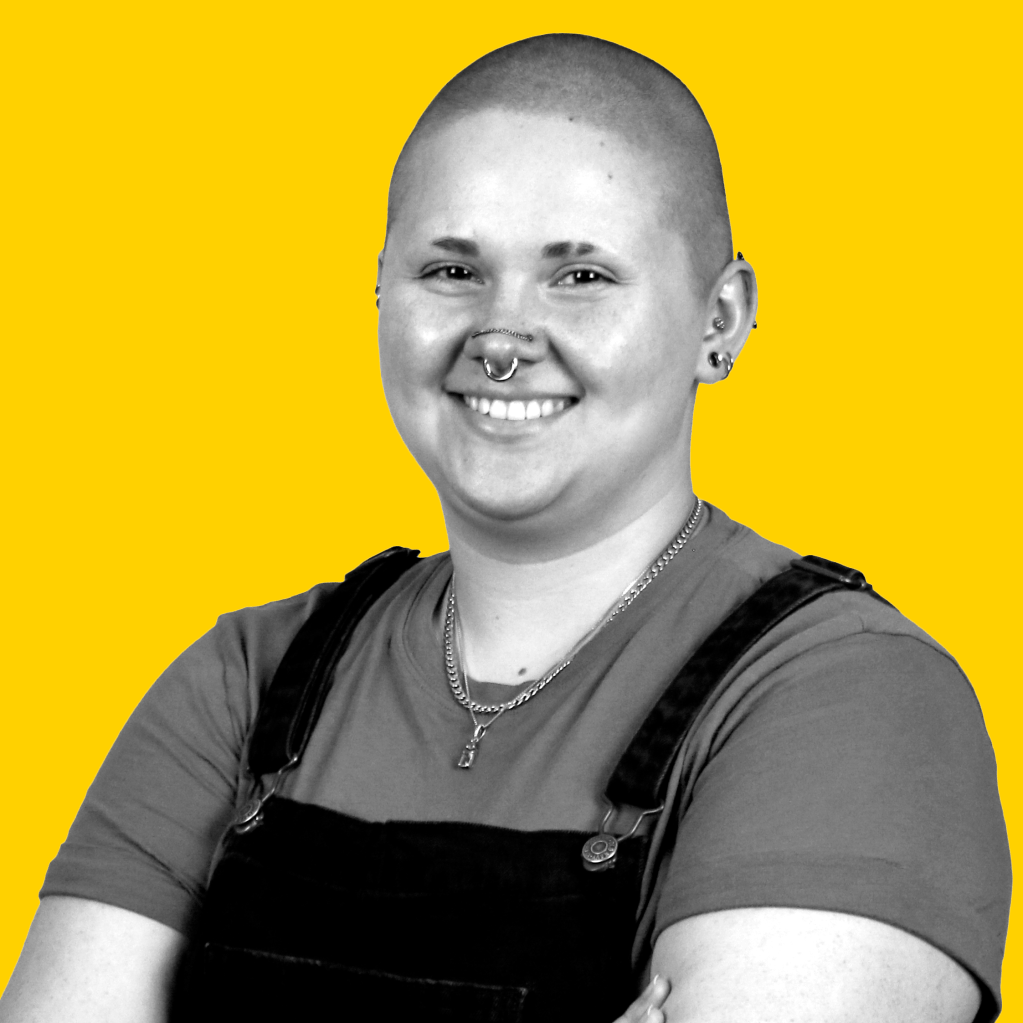 A photo of a bald, genderqueer individual with piercings, wearing overalls and a t-shirt.